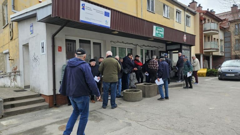 Grupa ludzi oczekuje na zewnątrz budynku, niekt&oacute;rzy trzymają dokumenty, w tle budynek mieszkalny.