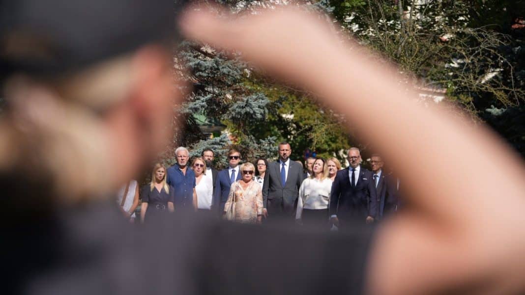 Grupa urzędnik&oacute;w i mieszkańc&oacute;w na zewnątrz, widziana przez osobę trzymającą telefon, w tle drzewa i budynki.