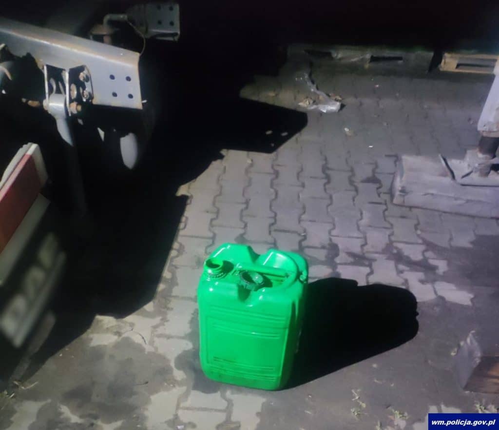 Zdjęcie przedstawia zielony kanister na tle podjazdu, obok samochodu i innych przedmiot&oacute;w, nocą.