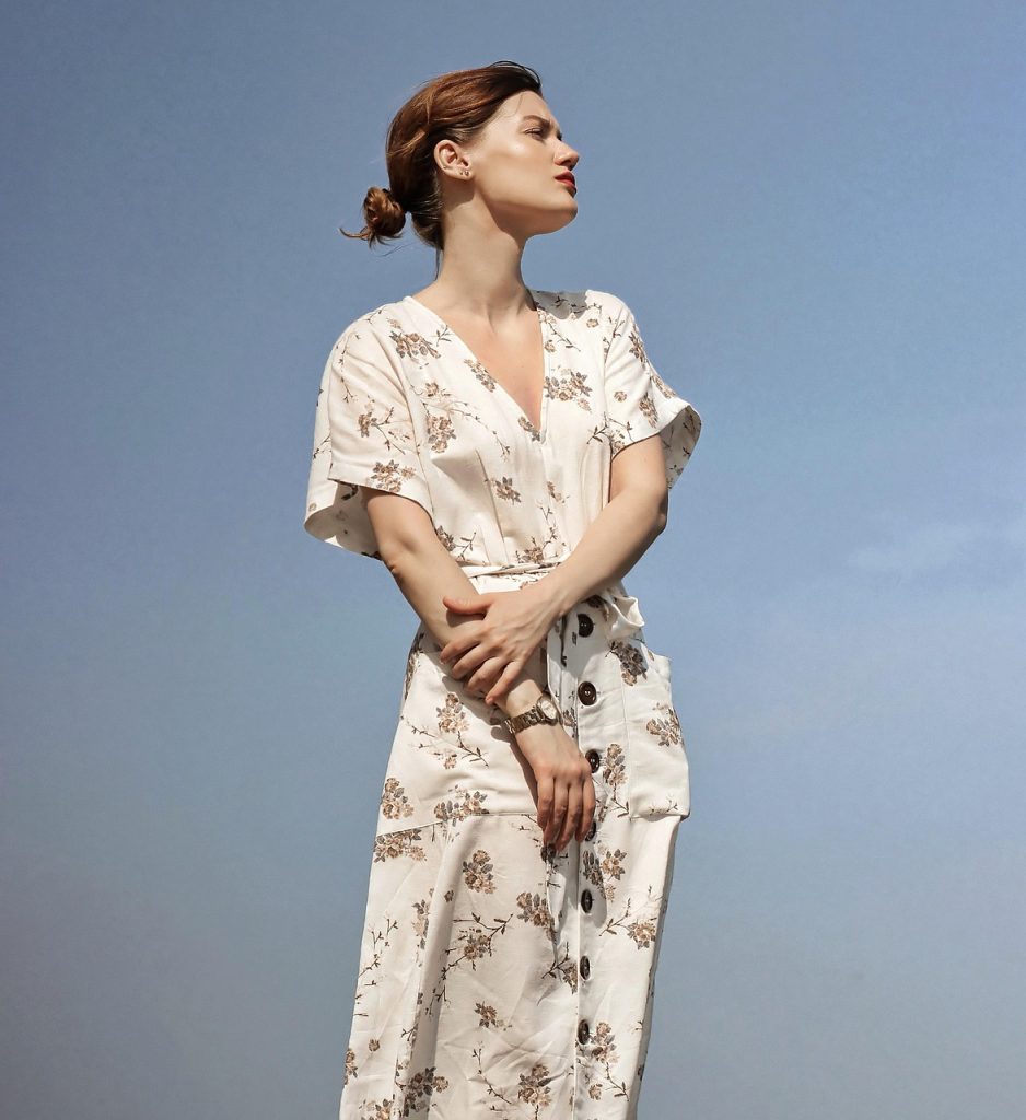 Kobieta w eleganckiej, jasnej sukience z kwiatowym wzorem na tle nieba.