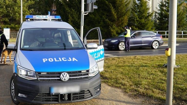 Policja na ulicy, funkcjonariusze rozmawiają i patrolują teren, w tle drzewa i znaki drogowe.
