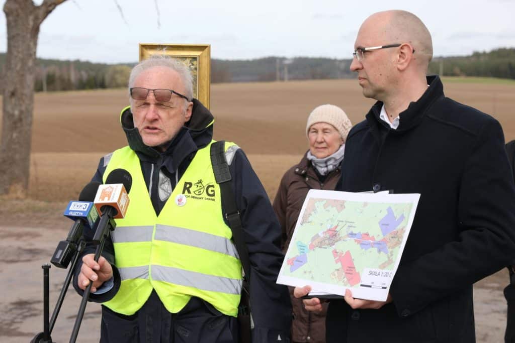 Mężczyzna w kamizelce odblaskowej i drugi z mapą na tle p&oacute;l i drzew, podczas konferencji na zewnątrz.