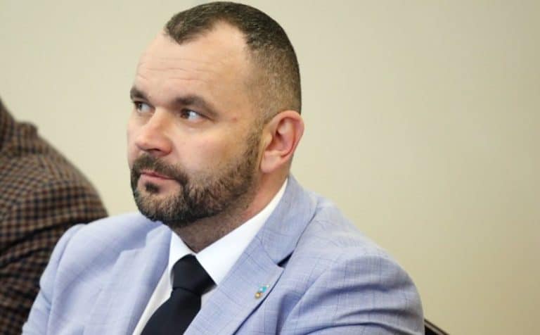 Mężczyzna w jasnoszarym garniturze i czarnej krawat, siedzi na spotkaniu, patrzy w bok.