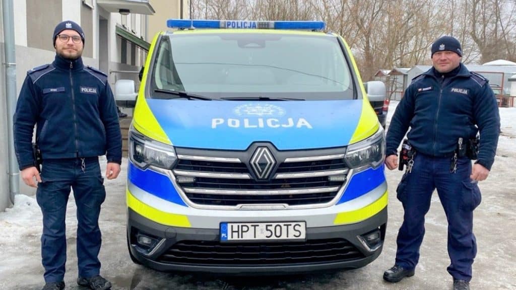 Dw&oacute;ch policjant&oacute;w stojących obok radiowozu na tle zimowego otoczenia, w tym śniegu i dom&oacute;w.