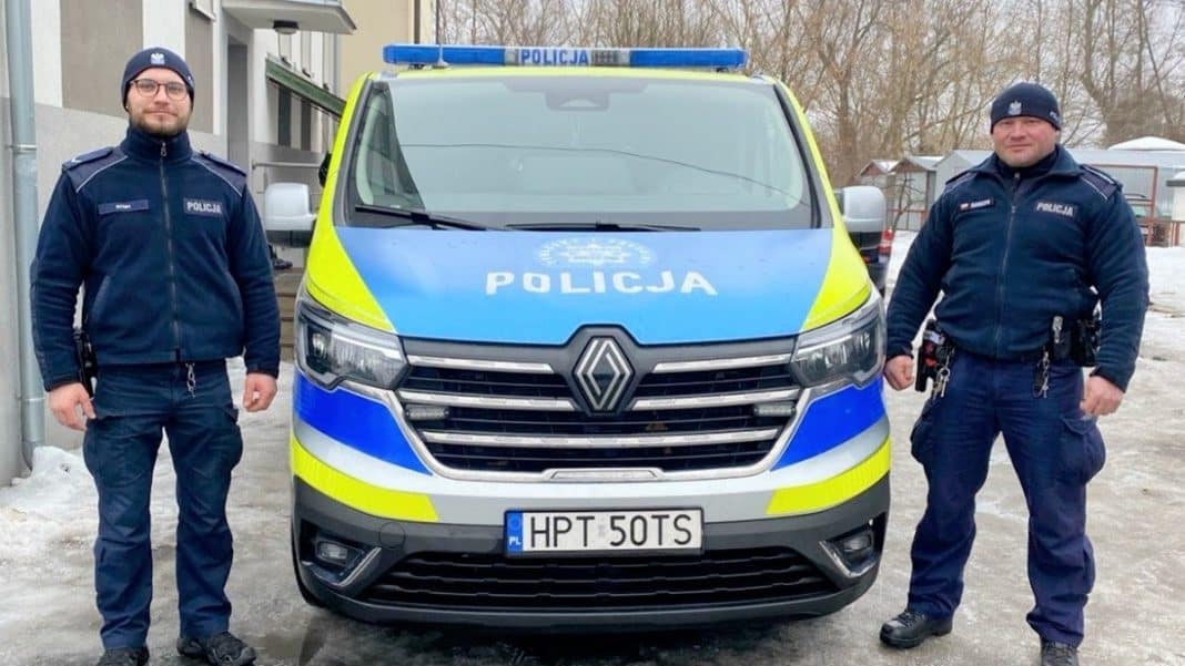 Dw&oacute;ch policjant&oacute;w stojących obok radiowozu na tle zimowego otoczenia, w tym śniegu i dom&oacute;w.