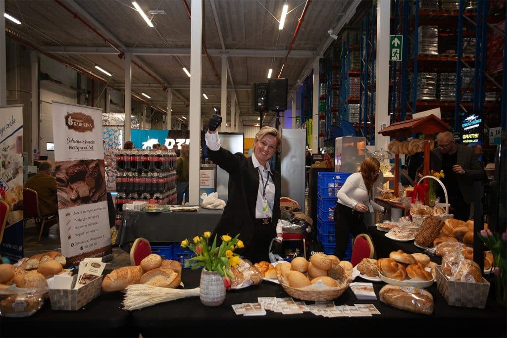 Stoisko piekarni z r&oacute;żnymi wypiekami i przedstawicielka uśmiechnięta na tle targ&oacute;w w hali przemysłowej.