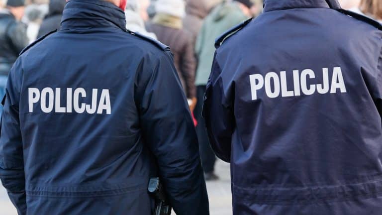 Dwoje policjant&oacute;w w uniformach na tle tłumu ludzi, widoczne tylne strony ich kurtek z napisem 'POLICJA'.