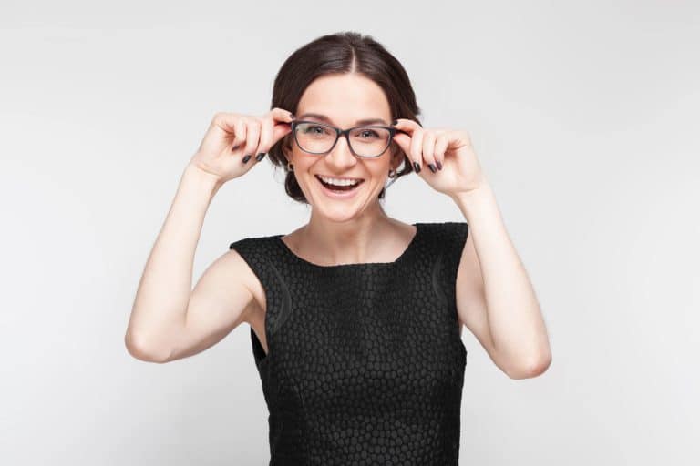 Uśmiechnięta kobieta w okularach, trzymająca je na nosie, na jasnym tle.
