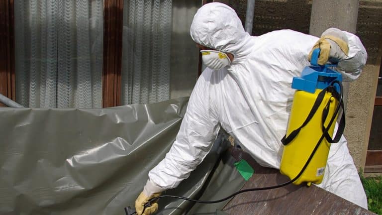 Pracownik w ochronnym kombinezonie i maseczce dezynfekuje teren wok&oacute;ł fermy drobiu.