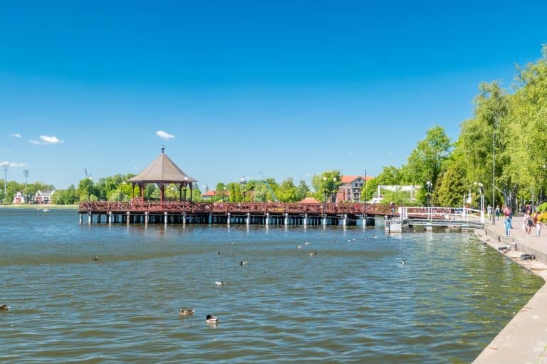 Widok na uszkodzony bulwar nad jeziorem z drewnianą kładką i zielonymi drzewami w tle.