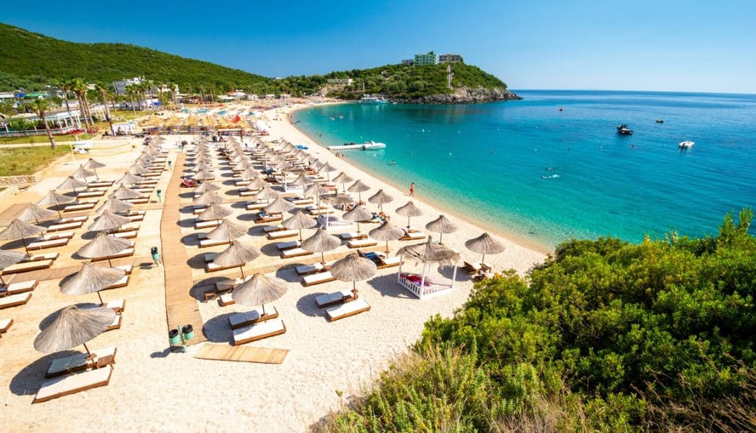 Plaża z leżakami i parasolami nad morzem, otoczona zielonymi wzg&oacute;rzami i błękitnym niebem.