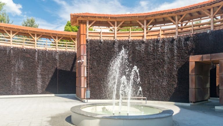 Widok na nowoczesny park z fontanną i drewnianą konstrukcją, w tle niebo z chmurami.