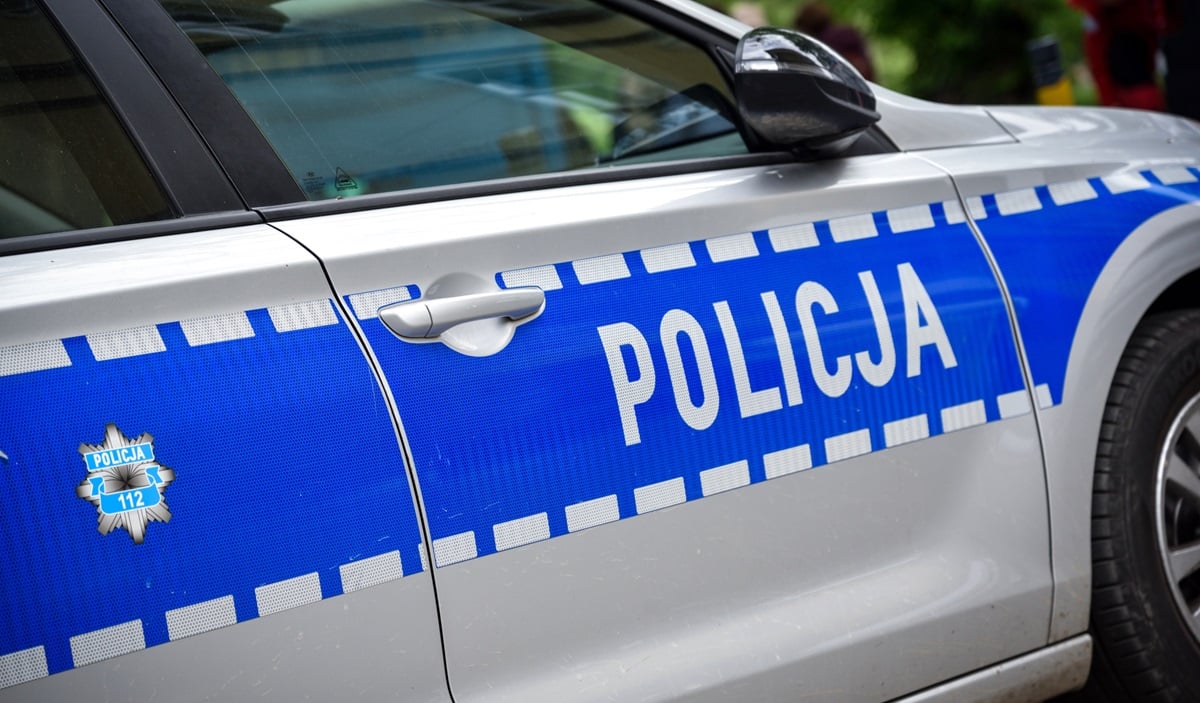 Samochód policyjny na ulicy, widoczny napis 'Policja' na drzwiach.