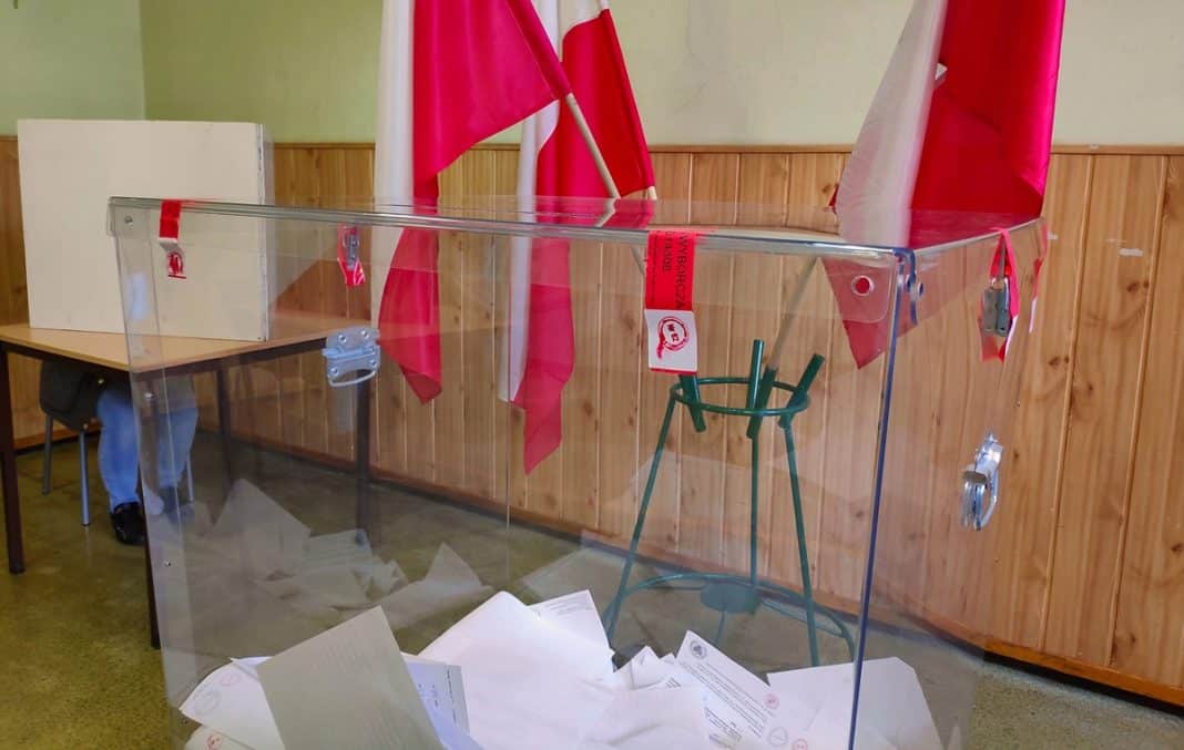 Przezroczysta urna wyborcza z dokumentami, za nią flagi na tle drewnianej ściany.