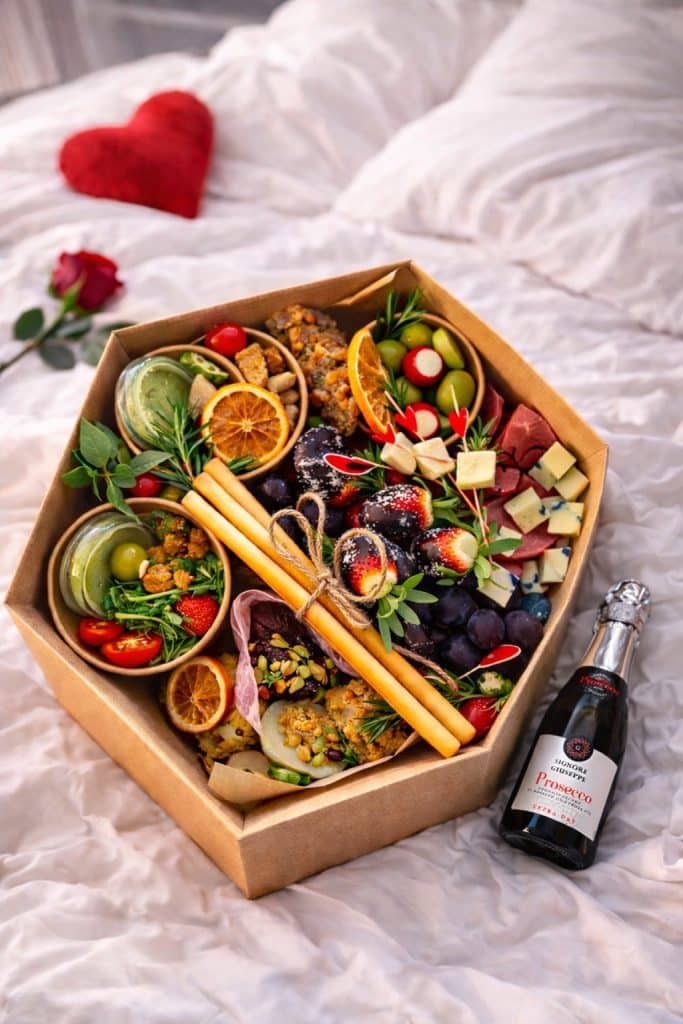 Zestaw przekąsek w boxie na imprezę, z owocami, serami i wędlinami, na tle ł&oacute;żka z sercem z czerwonego filcu.