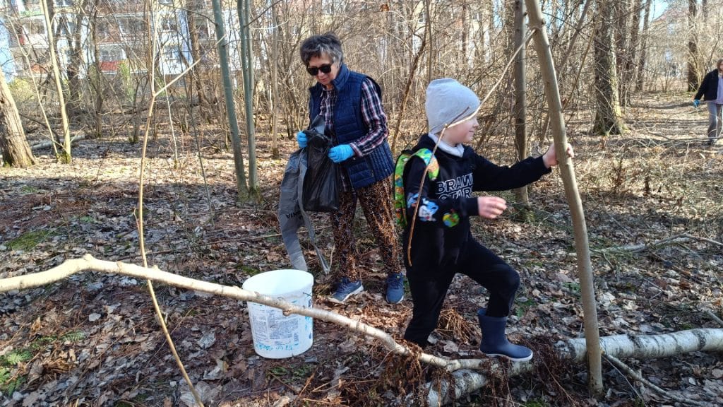 Mieszkańcy i dzieci sprzątają las w słoneczny dzień, zbierając odpady i porządkując teren.