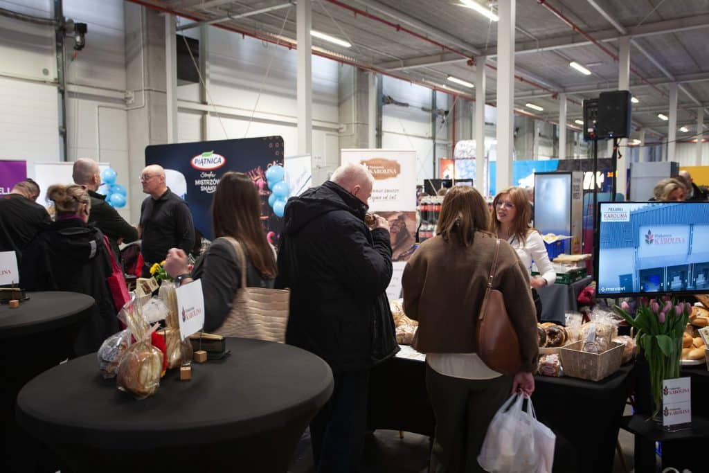 Tłum uczestnik&oacute;w na targach w hali przemysłowej, rozmawiają przy stoiskach z produktami spożywczymi i branżowymi.