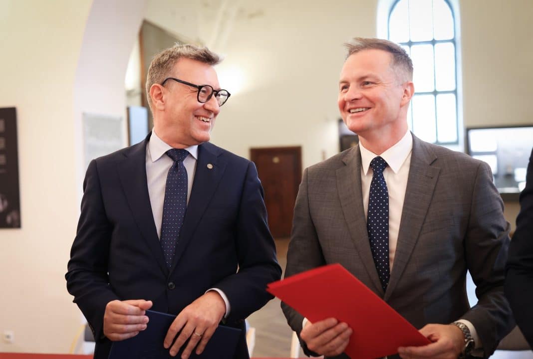 Dw&oacute;ch mężczyzn w garniturach rozmawia i uśmiecha się podczas spotkania w pomieszczeniu.