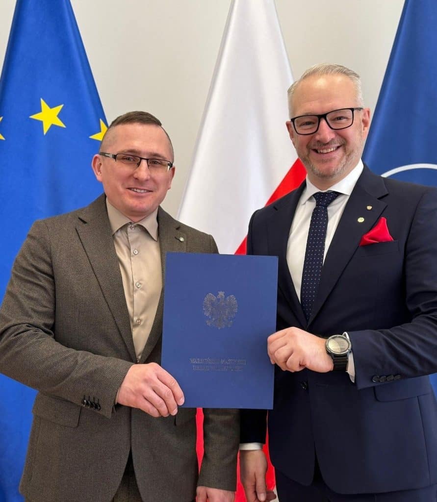 Dw&oacute;ch mężczyzn w garniturach trzymających dokument, za nimi flagi UE, Polski i NATO.