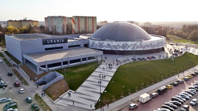 Widok na halę Urania w Olsztynie, otoczoną parkingiem i spacerującymi ludźmi, z wieloma blokami w tle.