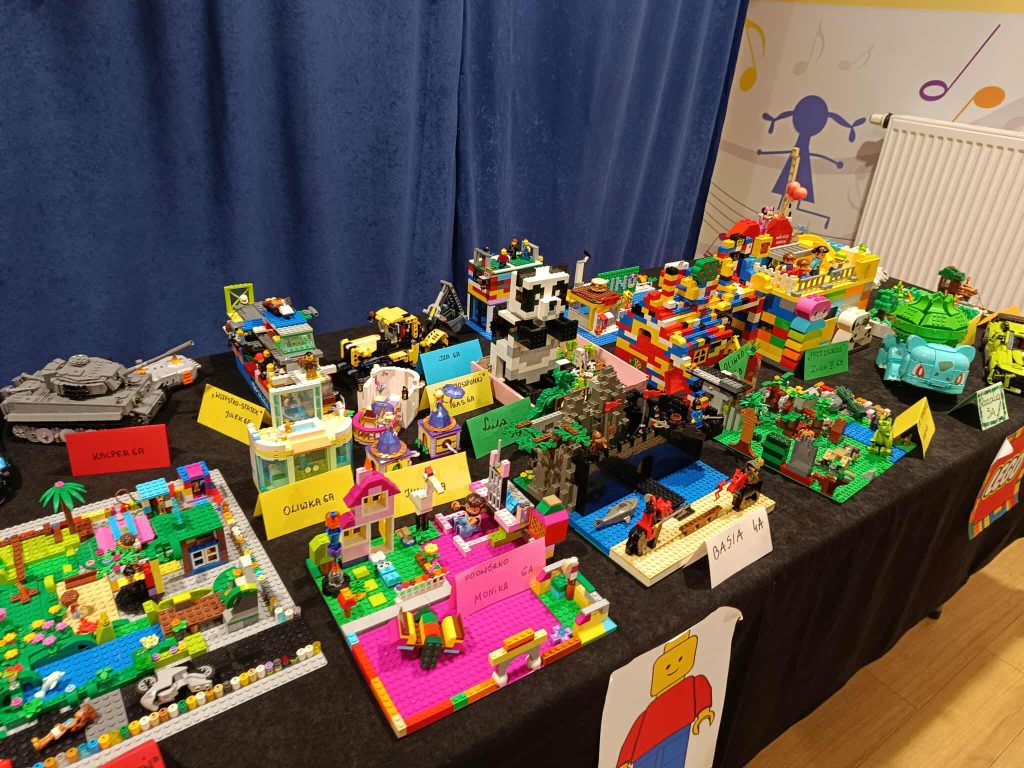 Na zdjęciu kolorowe modele z klock&oacute;w LEGO na stole, oznaczone etykietami, w tle ciemnoniebieska zasłona.