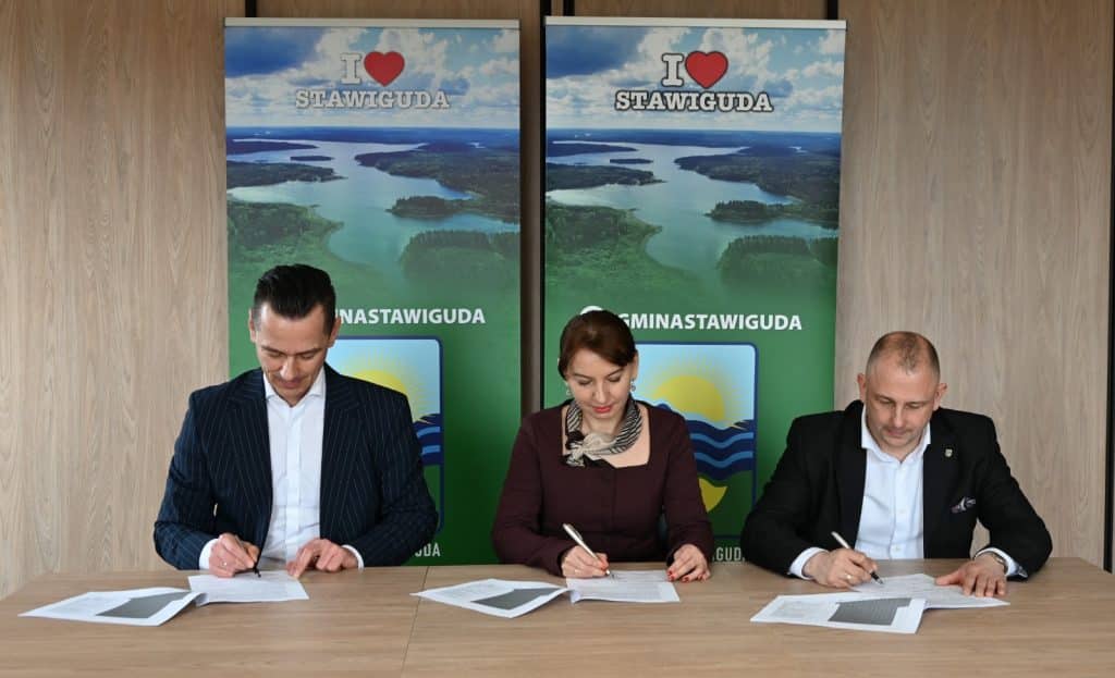 Trzy osoby podpisują umowę przy stołach, za nimi plakaty z nazwami gmin i krajobrazem jeziora.