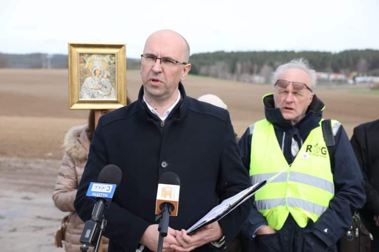 Mężczyzna przemawia podczas konferencji na tle pola, obok niego policjant w kamizelce odblaskowej.