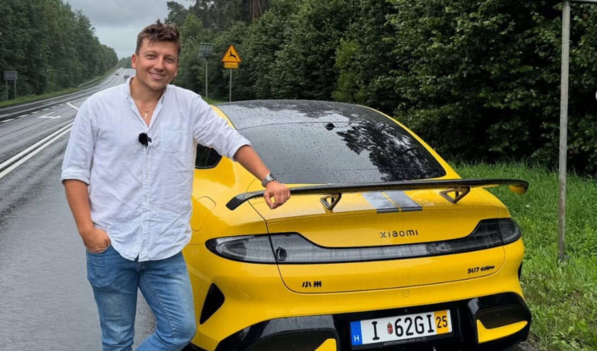 Mężczyzna stojący obok ż&oacute;łtego sportowego samochodu na deszczowej drodze, w tle zieleń i znaki drogowe.