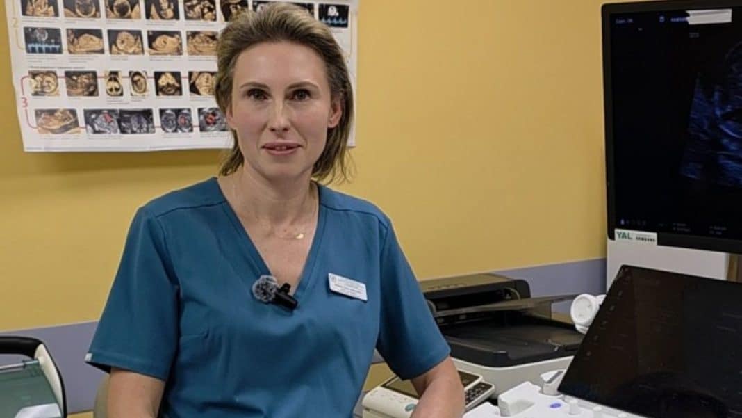 Lekarka w szpitalu przy komputerze, za nią plansza z obrazami prenatalnymi.