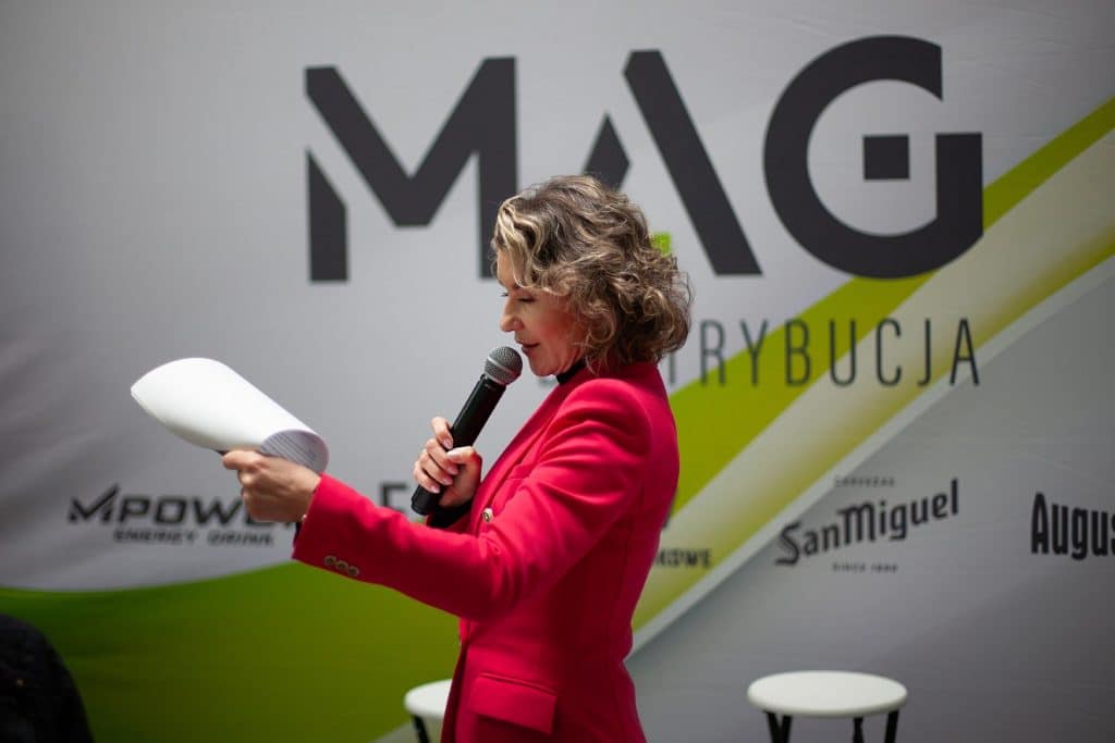 Kobieta w czerwonym garniturze przemawia do publiczności podczas targ&oacute;w w Gdańsku, za nią logo wydarzenia.