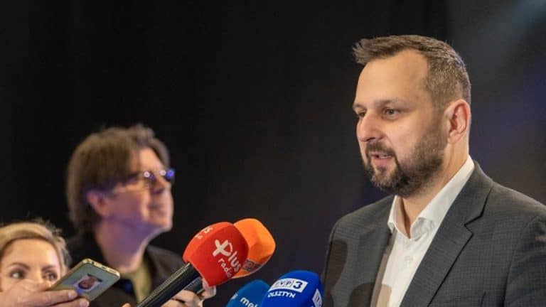 Mężczyzna w garniturze przemawia do medi&oacute;w podczas konferencji, obok mikrofony z logo r&oacute;żnych stacji.