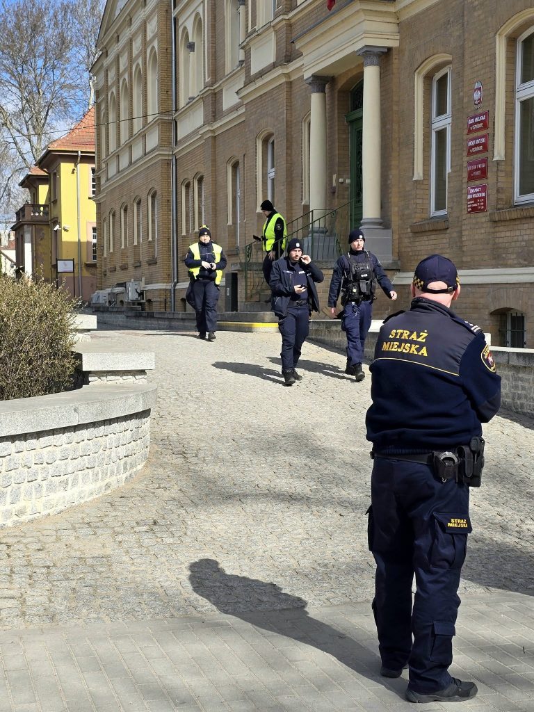 Grupa policjant&oacute;w i strażnik&oacute;w miejskich opuszcza budynek urzędu w Ostr&oacute;dzie po alarmie o ładunku wybuchowym.