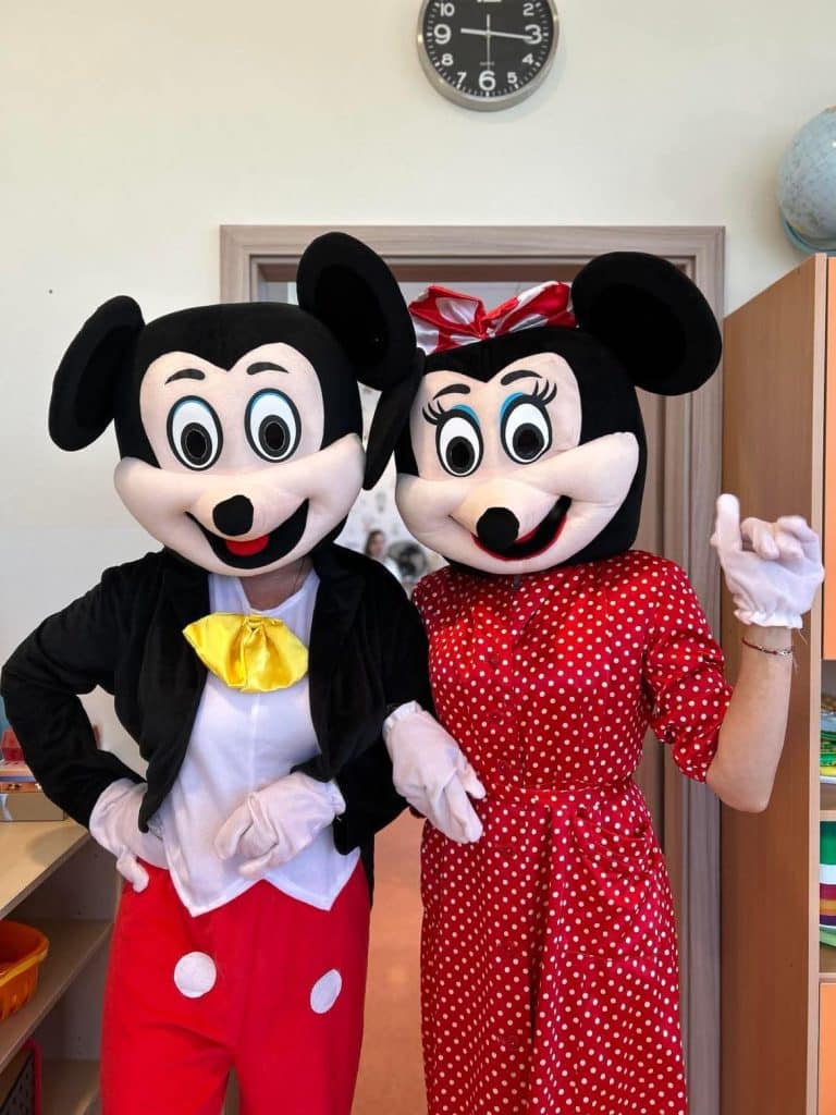 Dwie osoby w maskach postaci Myszki Miki i Minnie Mysz, ubrane w kolorowe stroje, w pomieszczeniu żłobka.