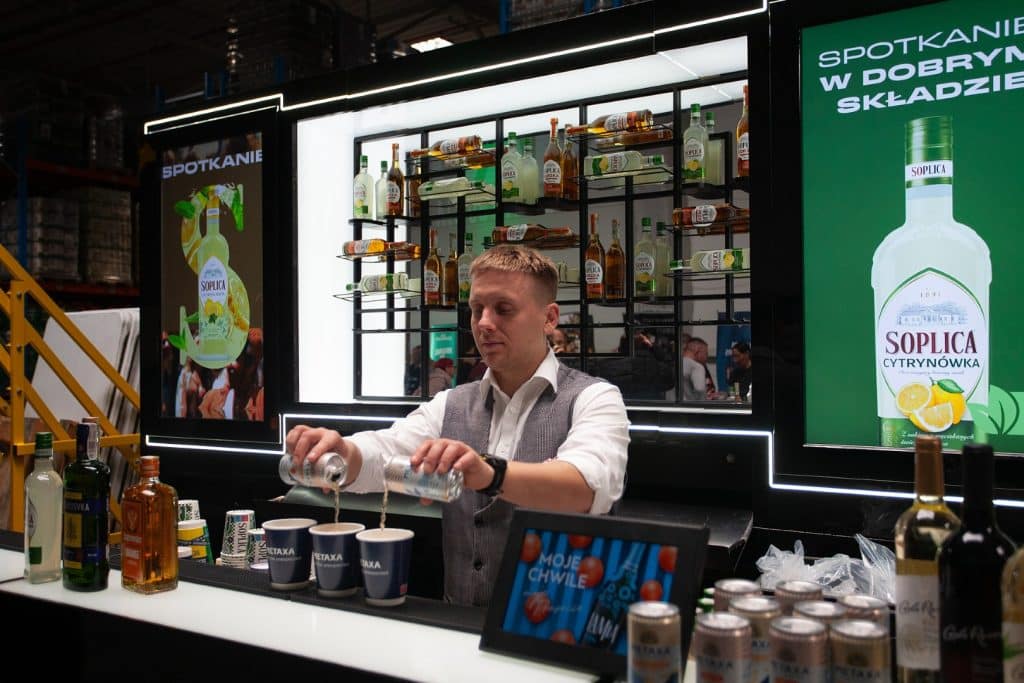 Przedstawiciel branży HoReCa serwuje napoje na targach w Gdańsku, za nim wyświetlają się reklamy produkt&oacute;w.