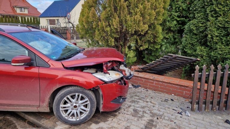 Uszkodzony czerwony samoch&oacute;d na chodniku, zniszczony płot i zarośla, po wypadku w Krotoszynach.