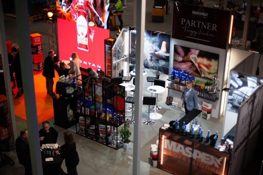 Stoisko targowe z produktami spożywczymi i napojami, z uczestnikami rozmawiającymi i prezentacjami.