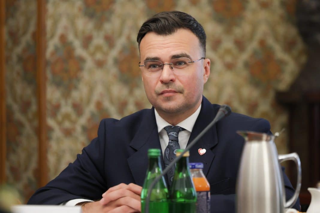 Mężczyzna w garniturze i okularach siedzi przy stole, za nim tapeta z kwiatowym wzorem.