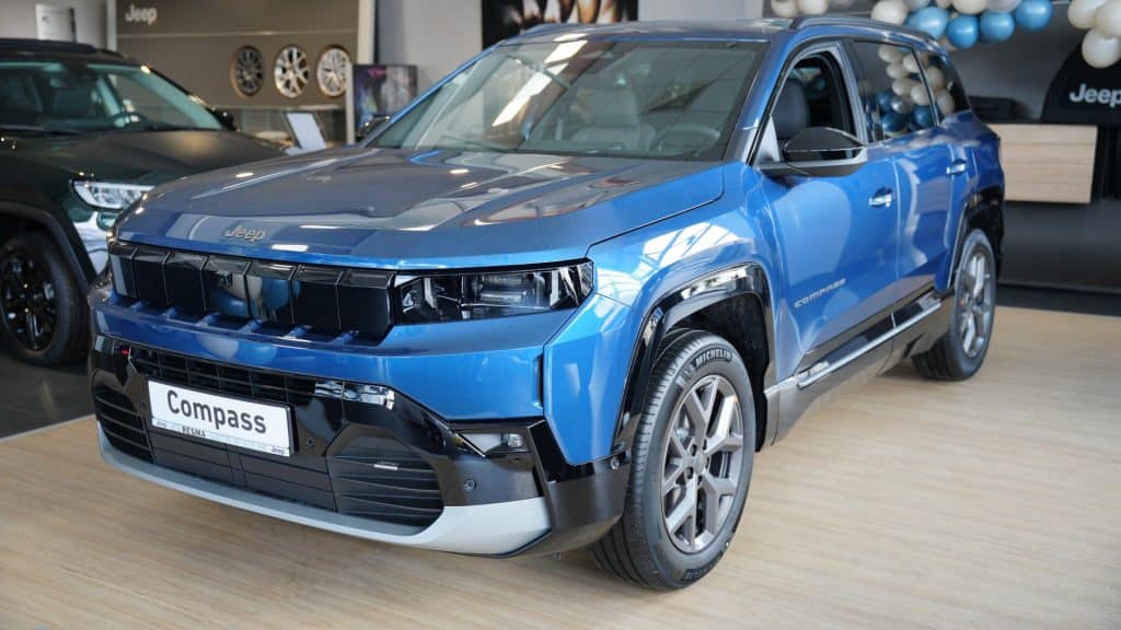 Nowy Jeep Compass w salonie samochodowym, niebieski, z nowoczesnym designem i dużym grillem.