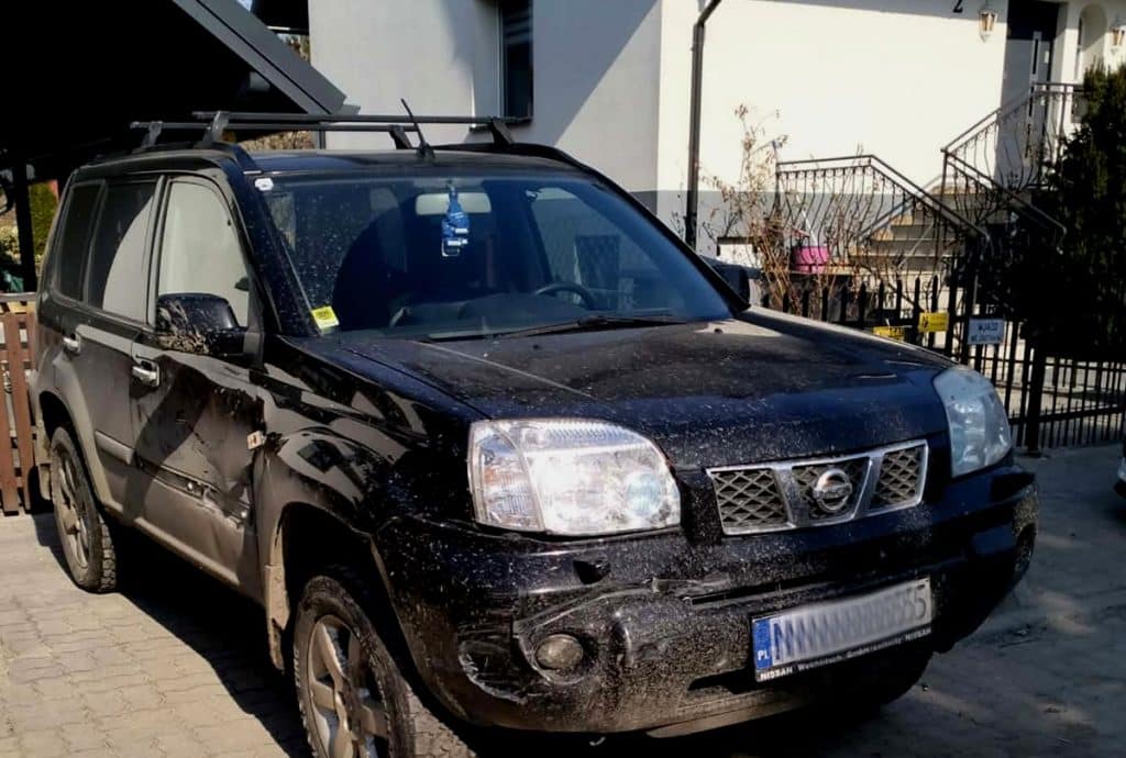 Uszkodzony czarny samoch&oacute;d na parkingu przy plaży, z widocznymi zarysowaniami i wgnieceniami po kolizji.