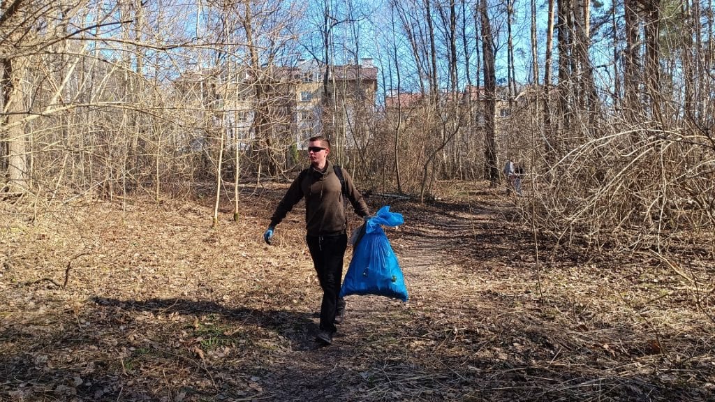 Mężczyzna z plecakiem i workiem na śmieci spaceruje po lesie w słoneczny dzień.