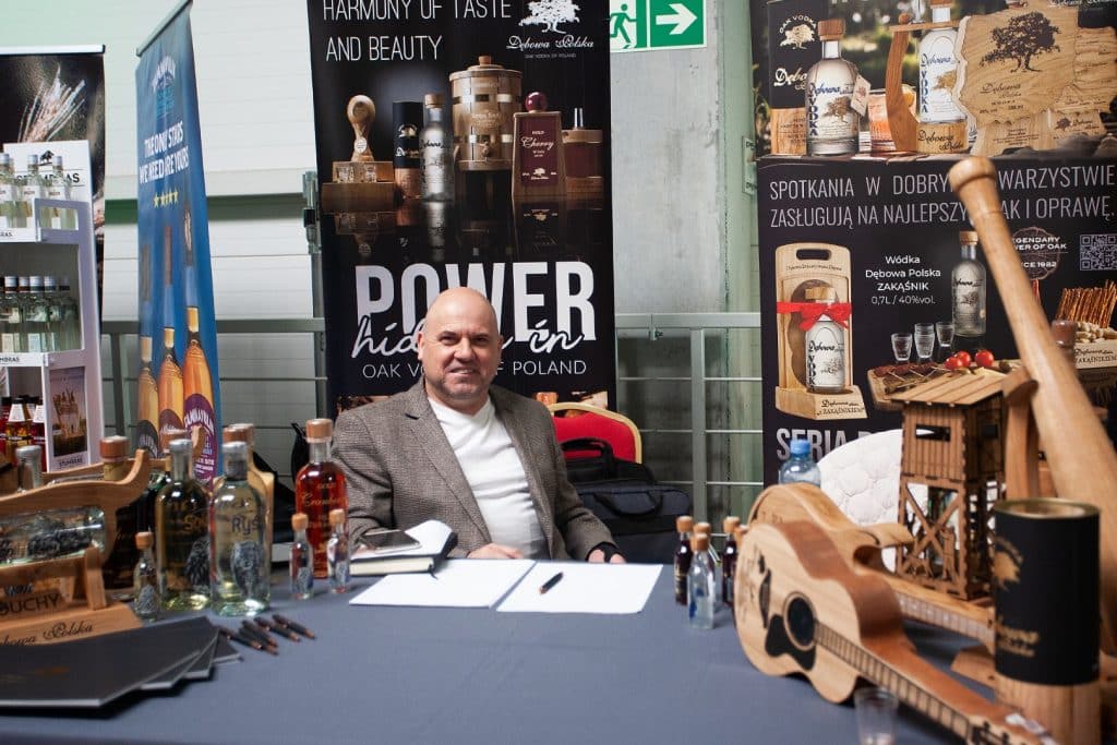 Mężczyzna siedzi przy stoisku targowym z produktami alkoholowymi i gadżetami, w tle banery promujące markę.
