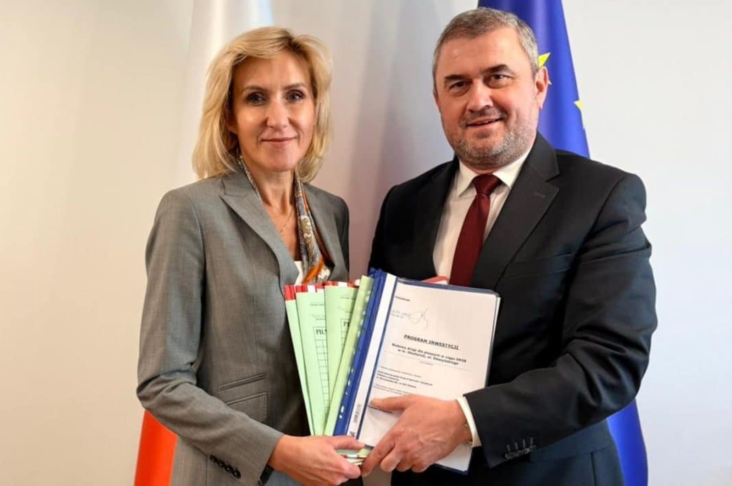 Dwoje os&oacute;b w formalnych ubraniach trzyma dokumenty, na tle flagi UE i biały ścian.