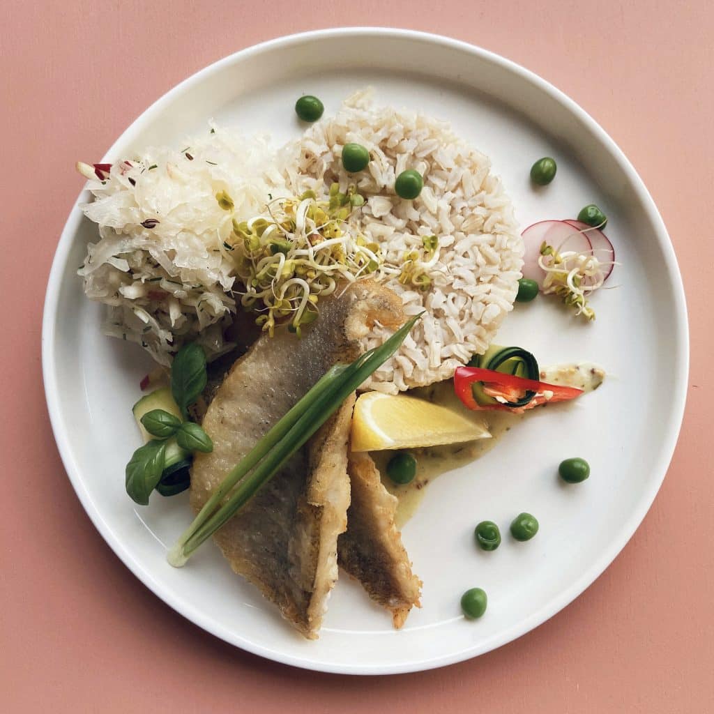 Zdjęcie zdrowego lunchu na białym talerzu, zawierającego rybę, ryż, warzywa i kiełki, na r&oacute;żowym tle.