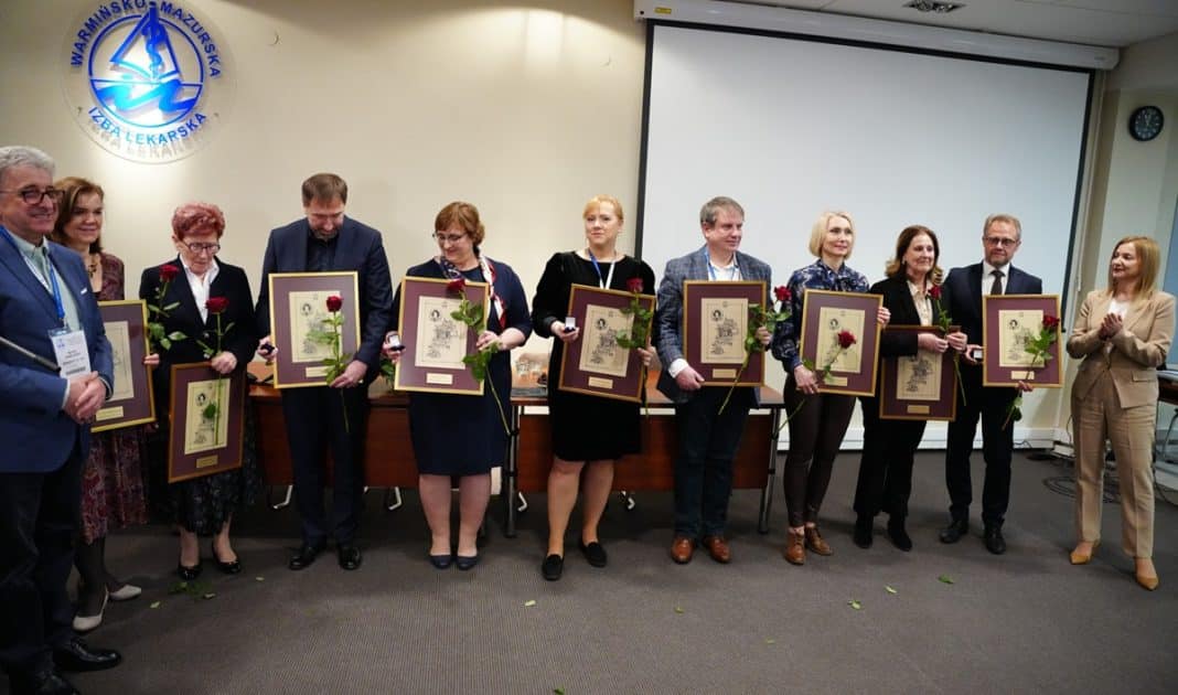 Grupa lekarzy z Warmii i Mazur podczas uroczystości wręczania wyr&oacute;żnień, trzymający ramki z odznaczeniami i kwiaty.