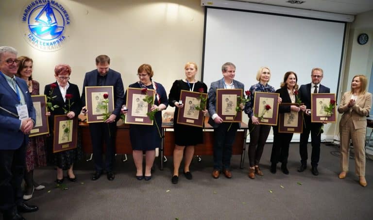 Grupa lekarzy z Warmii i Mazur podczas uroczystości wręczania wyr&oacute;żnień, trzymający ramki z odznaczeniami i kwiaty.