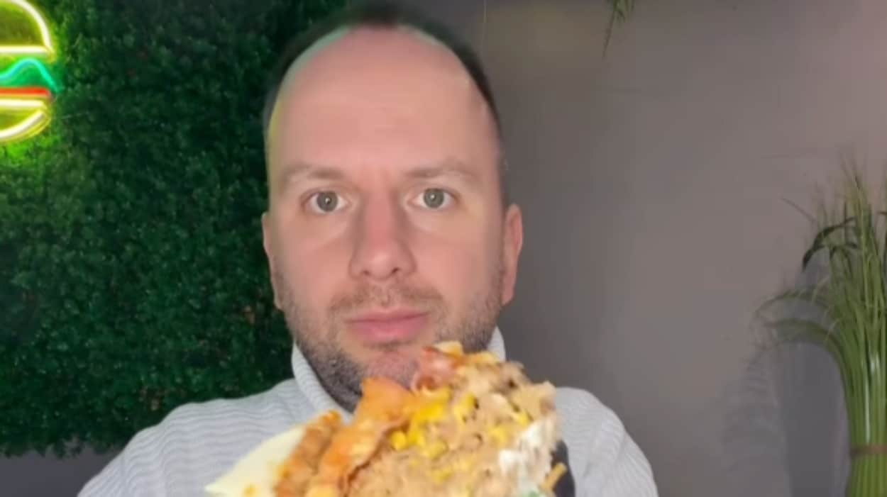 Mężczyzna trzymający burger w ręku, z wyrazem zadowolenia na twarzy, w tle zielona roślina i neonowe światło.