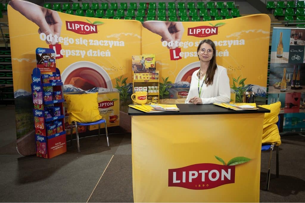 Stoiska promocyjne marki Lipton na targach w Olsztynie, kobieta przy stanowisku z materiałami reklamowymi.