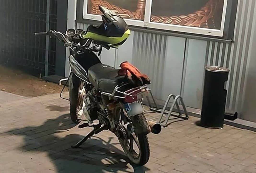 Motorower zaparkowany na chodniku, na tle metalowej ściany i okna, wieczorem.