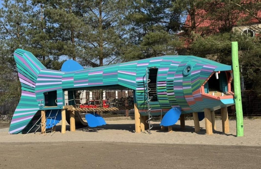 Kolorowa konstrukcja placu zabaw w kształcie ryby na tle drzew i budynk&oacute;w.