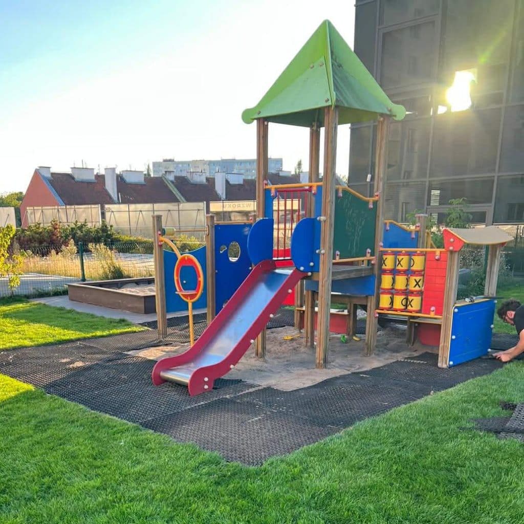 Plac zabaw na zewnątrz przedszkola z kolorową konstrukcją i zielonym dachem, otoczony zieloną trawą.